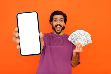 Foto de Préstamo en línea, crédito. Hombre joven indio guapo excitado en camiseta púrpura que muestra un gran teléfono celular con pantalla blanca en blanco y billetes en efectivo, aislado en el fondo colorido del estudio, maqueta - Imagen libre de derechos