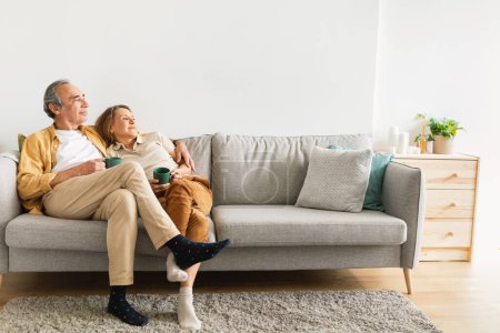 Photo for Happy long-lasting marriage. Senior husband and wife hugging, drinking coffee or tea resting on sofa, enjoying time together at home. Retirement lifestyle and happiness - Royalty Free Image