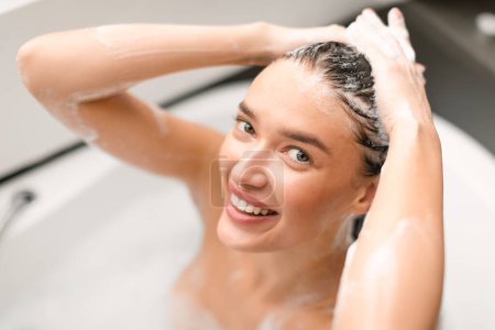 Téléchargez les photos : Attractive dame appliquant shampooing souriant à la caméra se laver la tête posant dans la baignoire dans la salle de bain à la maison. Bain de femme heureuse profiter des soins capillaires en utilisant des cosmétiques. Focus sélectif - en image libre de droit