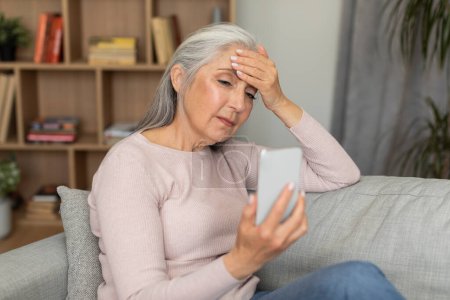 Photo for Sad tired caucasian old woman sits on sofa, looks at smartphone, presses head to forehead, suffers from migraine, pressure and stress in living room interior. App, bad news, health problems at home - Royalty Free Image