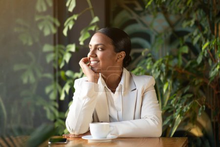 Foto de Alegre millennial mujer de negocios afroamericana en traje blanco mirar por la ventana en la oficina cafetería con el interior de plantas verdes. Negocios modernos, trabajo, coffee break, estilo de vida, anuncio y oferta - Imagen libre de derechos