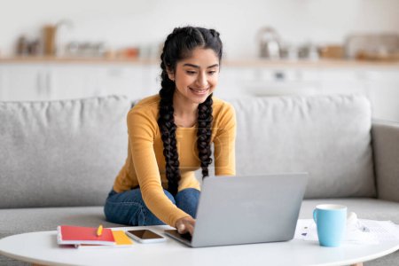 Foto de Educación en línea. Estudio femenino joven indio feliz con el ordenador portátil en casa, sonriendo señora estudiante oriental escribiendo en el teclado de la computadora mientras está sentado en el sofá en la sala de estar, disfrutando de la educación a distancia - Imagen libre de derechos