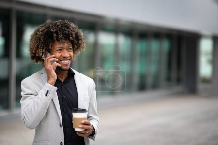 Téléchargez les photos : Heureux entrepreneur noir d'âge moyen parlant sur smartphone et buvant du café dans une tasse à emporter, marchant à l'extérieur près d'un immeuble de bureaux, copiez l'espace. Homme d'affaires utilisant gadget pour les affaires à distance - en image libre de droit