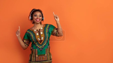 Téléchargez les photos : Joyeux joyeux millénaire dame noire en costume africain traditionnel lumineux en utilisant des écouteurs sans fil, écouter de la musique et de la danse, en regardant l'espace de copie pour la publicité sur fond de studio orange - en image libre de droit