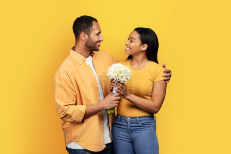 Photo for Happy millennial african american husband gives bouquet and hugs to wife, congratulates with birthday, anniversary isolated on yellow studio background. Relationship, romance, love and date - Royalty Free Image