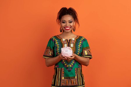 Téléchargez les photos : Économie, investissement, concept de dépôt. Joyeux jolie jeune femme noire en costume africain tenant tirelire et sourire, économiser de l'argent pour l'avenir, fond de studio orange, espace de copie - en image libre de droit
