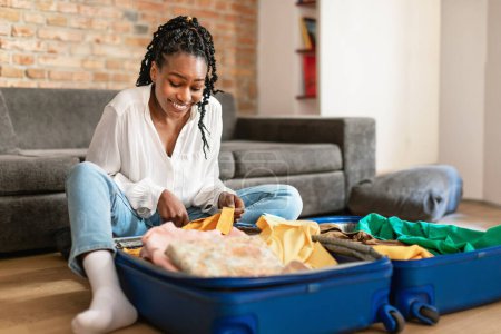 Photo for Tourism and traveling concept. Black lady packing suitcase and smiling, sitting on floor at home, free space. Happy female traveler preparing for journey - Royalty Free Image