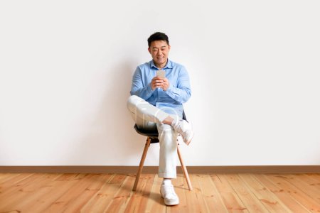 Foto de Hombre coreano de mediana edad utilizando el teléfono celular, mensajes de texto o navegar por Internet mientras está sentado en la silla contra el fondo blanco de la pared. Gadgets y concepto de comunicación móvil. Longitud completa - Imagen libre de derechos