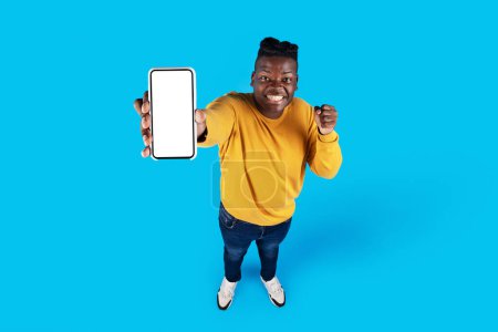 Téléchargez les photos : Homme noir excité démontrant Smartphone vierge avec écran blanc et célébrant le succès, homme afro-américain joyeux bénéficiant d'une nouvelle application mobile ou offre en ligne, debout sur fond bleu, Mockup - en image libre de droit