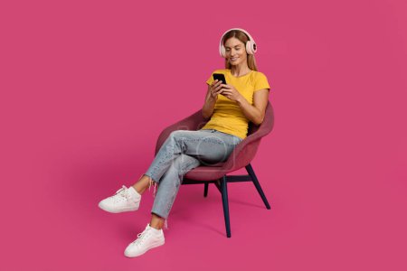 Photo for Happy middle aged european lady sit in armchair, typing on smartphone, listen music in wireless headphones isolated on pink studio background. Watch video on device, online call and audio app - Royalty Free Image