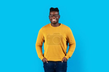 Photo for Smiling Young Black Man Wearing Stylish Eyeglasses Posing Over Blue Studio Background, Handsome Millennial African American Guy Keeping Hands In Pockets And Looking At Camera, Copy Space - Royalty Free Image
