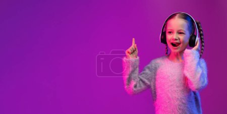 Téléchargez les photos : Joyeux enfant jolie préadolescente aux cheveux blonds en utilisant de nouveaux écouteurs sans fil blancs, pointant vers l'espace de copie et souriant sur fond de studio rose dans la lumière au néon, écoutant de la musique, bannière web - en image libre de droit