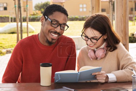 Photo for Smiling millennial black man and european lady modern students read books, study together at university campus, outdoor. Knowledge, education, test preparation and coffee break - Royalty Free Image