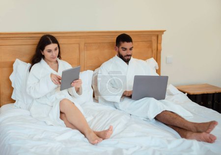 Téléchargez les photos : Couple freelancers utilisant ordinateur portable et tablettes numériques travaillant en ligne et naviguant sur Internet assis dans le lit à la maison, portant des peignoirs blancs. Concept de technologie et gadgets - en image libre de droit