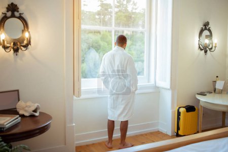 Photo for Back View Of Man Wearing White Bathrobe Standing Near Window In Luxury Hotel Suite Indoor. Unrecognizable Male Traveler Relaxing In Resort Enjoying Vacation. Full Length Shot - Royalty Free Image