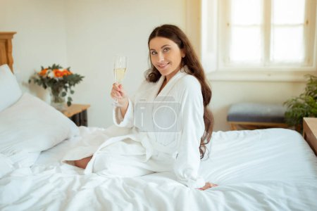 Téléchargez les photos : Happy Woman tenant un verre de vin mousseux appréciant boire de l'alcool relaxant assis sur le lit portant un peignoir blanc dans une suite hôtelière de luxe à l'intérieur, souriant à la caméra. Concept de vacances - en image libre de droit