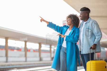 Foto de Pareja negra de pie en la plataforma en la estación de tren y señalando lejos, jóvenes cónyuges afroamericanos felices esperando la llegada del tren en el centro de tren, disfrutando de viajar juntos, Copiar el espacio - Imagen libre de derechos
