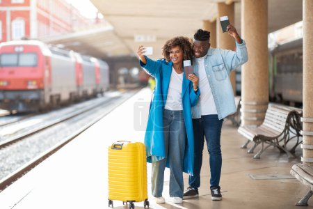 Téléchargez les photos : Voyage de vacances. Joyeux conjoints noirs prenant Selfie à la gare, heureux couple afro-américain capturant des photos avec Smartphone en attendant le train, tenant passeport et billets - en image libre de droit