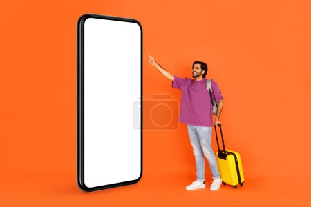 Photo for Cheerful handsome young indian man tourist standing by big smartphone with white empty display, touching blank screen, isolated on orange studio background, online check-in, flights booking, mockup - Royalty Free Image