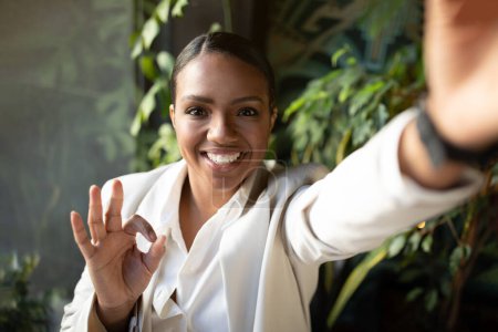 Photo for Positive millennial african american lady in white suit doing ok hand gesture, taking selfie in cafe office with green plants interior. Video call, business blog, communication, app recommendation - Royalty Free Image