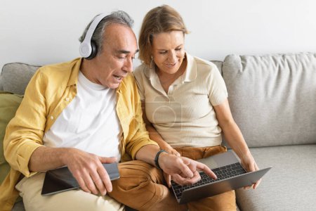 Photo for Two senior people share laptop looking and planning new travel, surfing internet, web surfing with lcomputer and headphones, sitting on sofa at home, copy space - Royalty Free Image