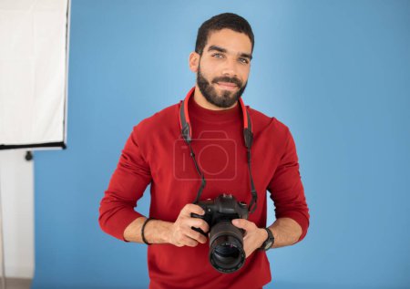 Téléchargez les photos : Photographe professionnel heureux beau jeune homme hispanique barbu en tenue décontractée posant avec appareil photo numérique dans un studio photo avec toile de fond bleue, espace de copie. Emploi et profession concept - en image libre de droit