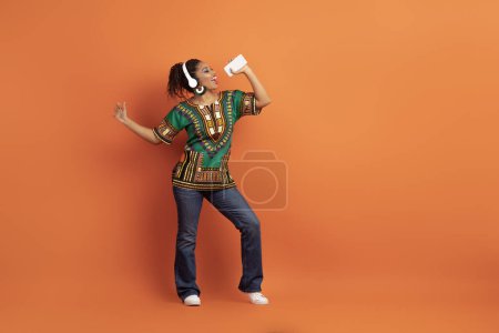 Téléchargez les photos : Joyeux jouet jeune femme africaine heureuse portant des vêtements nationaux chantant karaoké, en utilisant des écouteurs sans fil et smartphone comme microphone, danse, fond orange, espace de copie, pleine longueur - en image libre de droit