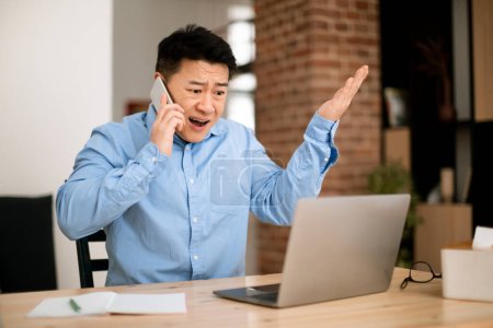 Téléchargez les photos : Émotionnel en colère asiatique moyen âge homme d'affaires travaillant de la maison, ayant conversation téléphonique, regardant l'écran d'ordinateur portable et gestuelle, copier l'espace - en image libre de droit