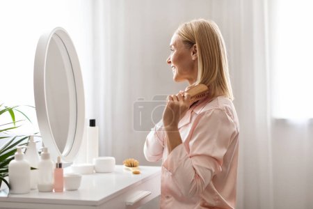 Photo for Beautiful Middle Aged Woman Brushing Her Blonde Hair With Comb While Sitting At Dressing Table In Bedroom At Home, Happy Mature Female Looking At Her Reflection And Smiling, Side View - Royalty Free Image