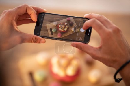 Photo for Cropped of male hands holding smartphone, food blogger influencer or pastry chef taking photo of sweets diverse cakes, plate with pastry on wooden table, selective focus on cell phone screen - Royalty Free Image
