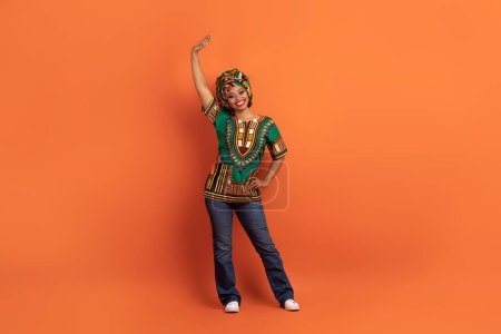 Téléchargez les photos : Positif souriant joyeux jeune jolie femme noire en costume africain traditionnel agitant signe de bonjour geste sur fond de studio orange, studio pleine longueur, espace de copie - en image libre de droit