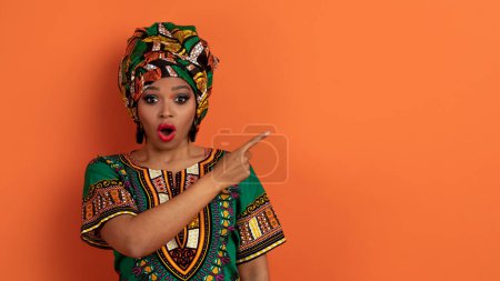 Photo for Unbelievable offer. Shocked sirprised beautiful young black woman in colorful traditional african clothes showing copy space for advertisement, isolated on orange studio background, panorama - Royalty Free Image
