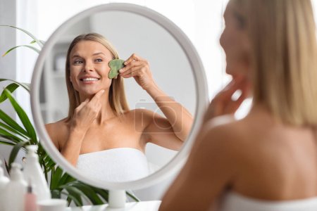 Hermosa mujer de mediana edad haciendo masaje facial con herramienta gua sha mientras está sentada cerca del espejo en casa, atractiva señora madura haciendo tratamientos de belleza después del baño, enfoque selectivo en la reflexión