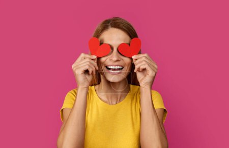 Photo for Laughing funny middle aged european blonde lady puts hearts to her eyes, like glasses, has fun, isolated on pink studio background. Valentine day, love, romance and lifestyle - Royalty Free Image