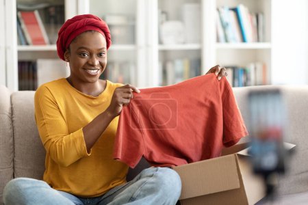 Photo for Female influencer stylish cheerful young black woman reviewing product. Excited african american millennial lady vlogging about unpacking good clothes and filming herself at home on cell phone - Royalty Free Image