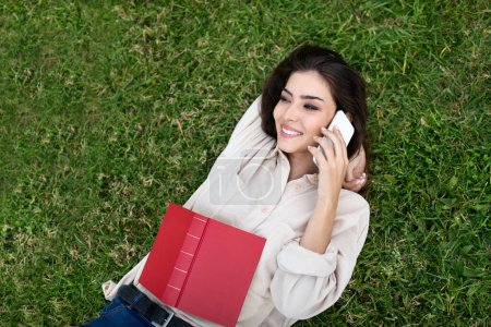 Photo for Cheerful millennial mixed race female student lies on grass with book, calls by phone in university campus, outdoor. Good news, gossip, break from study, knowledge, education, lifestyle in park - Royalty Free Image