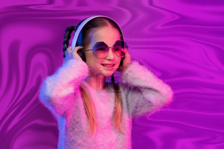 Foto de Retrato de dulce linda niña rubia linda en edad escolar en gafas de sol con auriculares inalámbricos modernos, niño feliz escuchando música, divertirse sobre fondo de lámina olográfica iridiscente rosa - Imagen libre de derechos