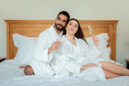 Photo for Romantic Honeymoon. Mixed Couple Holding Champagne Flutes Drinking Sparkling Wine Hugging Sitting In Bed Wearing Bathrobes In Luxury Hotel Suite Indoor. Vacation Accomodation Offer - Royalty Free Image
