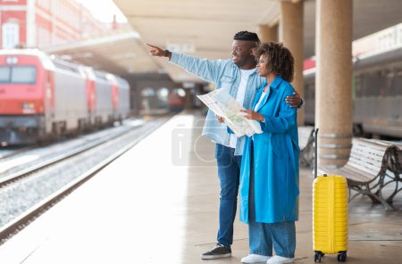 Téléchargez les photos : Par là. Couple de voyageurs noirs vérifiant l'itinéraire sur la carte de la ville tout en se tenant sur la gare après l'arrivée du train, jeune homme et femme afro-américain prévoyant où aller, copier l'espace - en image libre de droit