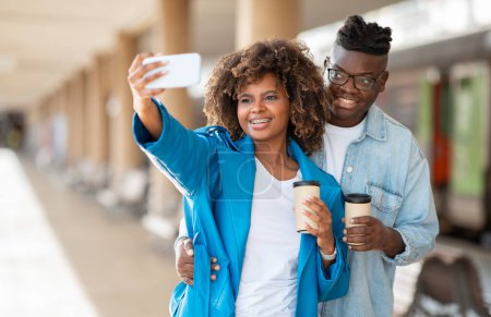 Foto de Alegre pareja negra tomando selfie en el teléfono inteligente mientras espera tren en la estación de tren, Feliz joven afroamericano hombre y mujer sosteniendo el café para llevar y sonriendo en la cámara, Espacio Copiar - Imagen libre de derechos