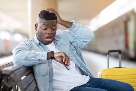 Téléchargez les photos : Homme noir stressé tard pour le train, jeune Afro-Américain bouleversé assis sur le banc à la gare et de vérifier le temps sur la montre-bracelet, Souffrant des problèmes tout en voyageant, Gros plan - en image libre de droit