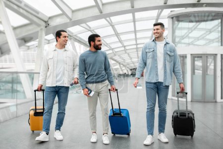 Foto de Tres turistas positivos hombres caminando con maletas de viaje sosteniendo pasaportes en la moderna terminal del aeropuerto interior. Tiro de longitud completa del grupo de pasajeros masculinos en la estación. Vacaciones y transporte - Imagen libre de derechos