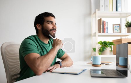 Téléchargez les photos : Heureux gars arabe millénaire coûteux avec barbe pense, prend des notes, travaille avec un ordinateur portable sur le lieu de travail dans l'intérieur de la salle de bureau. Entreprise, remue-méninges, idée créative, freelance et appel vidéo à la maison - en image libre de droit