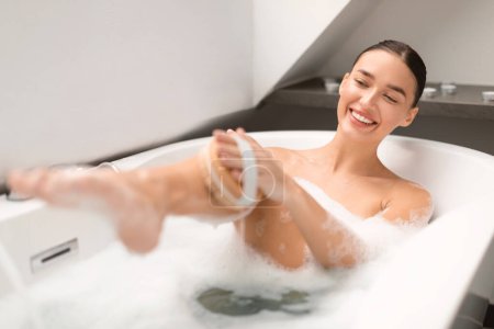 Téléchargez les photos : Soins du corps. Attrayant femme lavage et massage jambe avec brosse en bois baignade assis dans l'eau avec de la mousse dans la salle de bain moderne à l'intérieur. Soins de la peau et soins du corps Routine. Focus sélectif - en image libre de droit