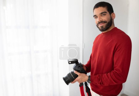 Téléchargez les photos : Positif joyeux attrayant hispanique gars en tenue décontractée élégant tenant appareil photo numérique et souriant, photographe professionnel appréciant son travail au studio de photo, espace de copie - en image libre de droit