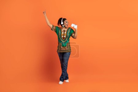 Téléchargez les photos : Joyeux positif insouciante jeune femme noire en vêtements africains chantant et dansant sur fond de studio orange, en utilisant des écouteurs sans fil et un téléphone portable comme micro, espace de copie, pleine longueur - en image libre de droit
