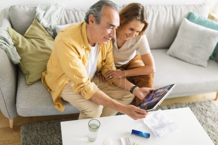 Photo for Happy senior couple smiling happily while having video call with family on digital tablet, sitting on sofa at home. Cheerful spouses enjoying their retirement - Royalty Free Image