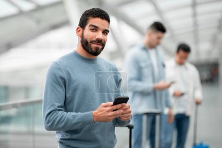 Téléchargez les photos : Arabe mâle passager textos sur téléphone portable dans l'aéroport moderne intérieur. Voyageur Guy utilisant Smartphone sourire à la caméra communiquant en ligne debout dans le terminal de départ. Publicité App Voyage - en image libre de droit