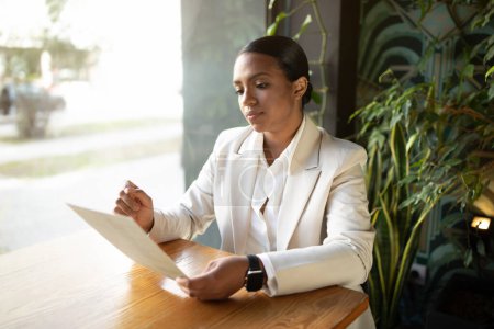 Foto de Graves concentrado millennial mujer de negocios afroamericana en traje blanco documento de lectura en la oficina de la cafetería con el interior de plantas verdes. Startup, negocios modernos, trabajo y estilo de vida - Imagen libre de derechos