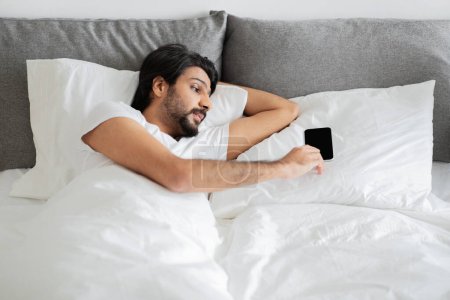 Photo for Serious young arab man in white t-shirt sleeps, wakes up, lies on bed, use smartphone with blank screen in bedroom interior. Rest with gadget, gadget addiction, morning news at home - Royalty Free Image